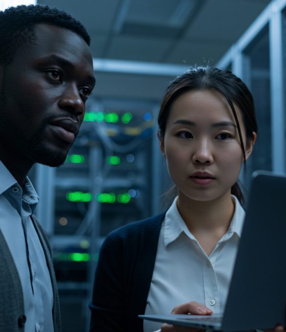 In a brightly lit data center, an African-American man and an Asian woman, both dressed in business attire, intently examine the network server status using a laptop. The focused professionals are reviewing server racks with blinking LED lights in this technology infrastructure workspace.